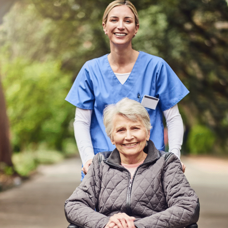 A caregiver standing behind an older woman seated in a wheelchair on a garden path.