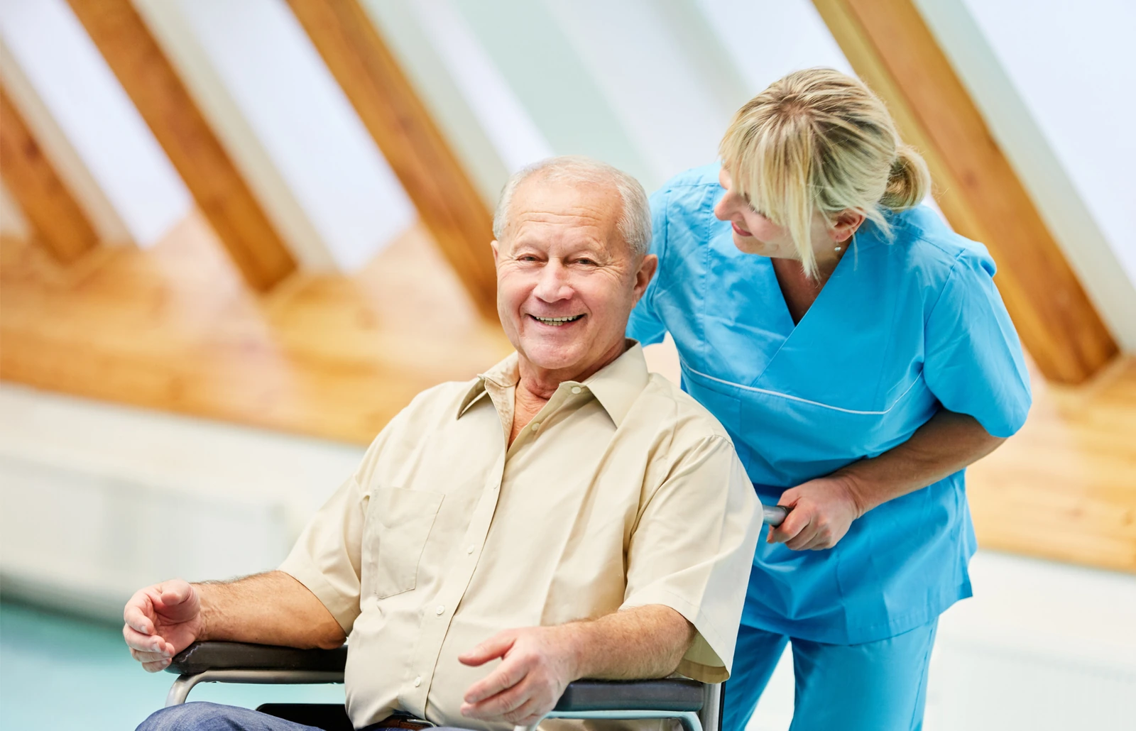 A care professional in turquoise scrubs speaks with an older man using a wheelchair. BestCareMatch editorial photo.