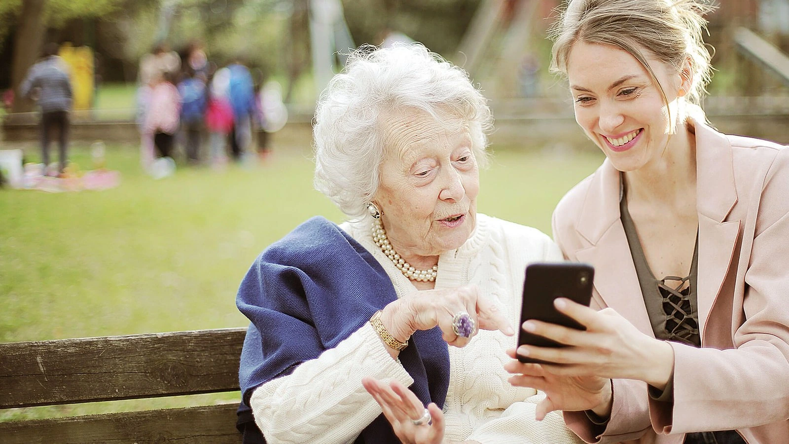An older woman and a younger woman look at a phone on a park bench. BestCareMatch editorial photo.