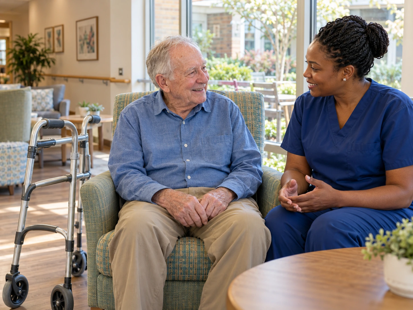 An older man talks with a care professional in blue scrubs, with a walker beside his chair. BestCareMatch illustrative photo.